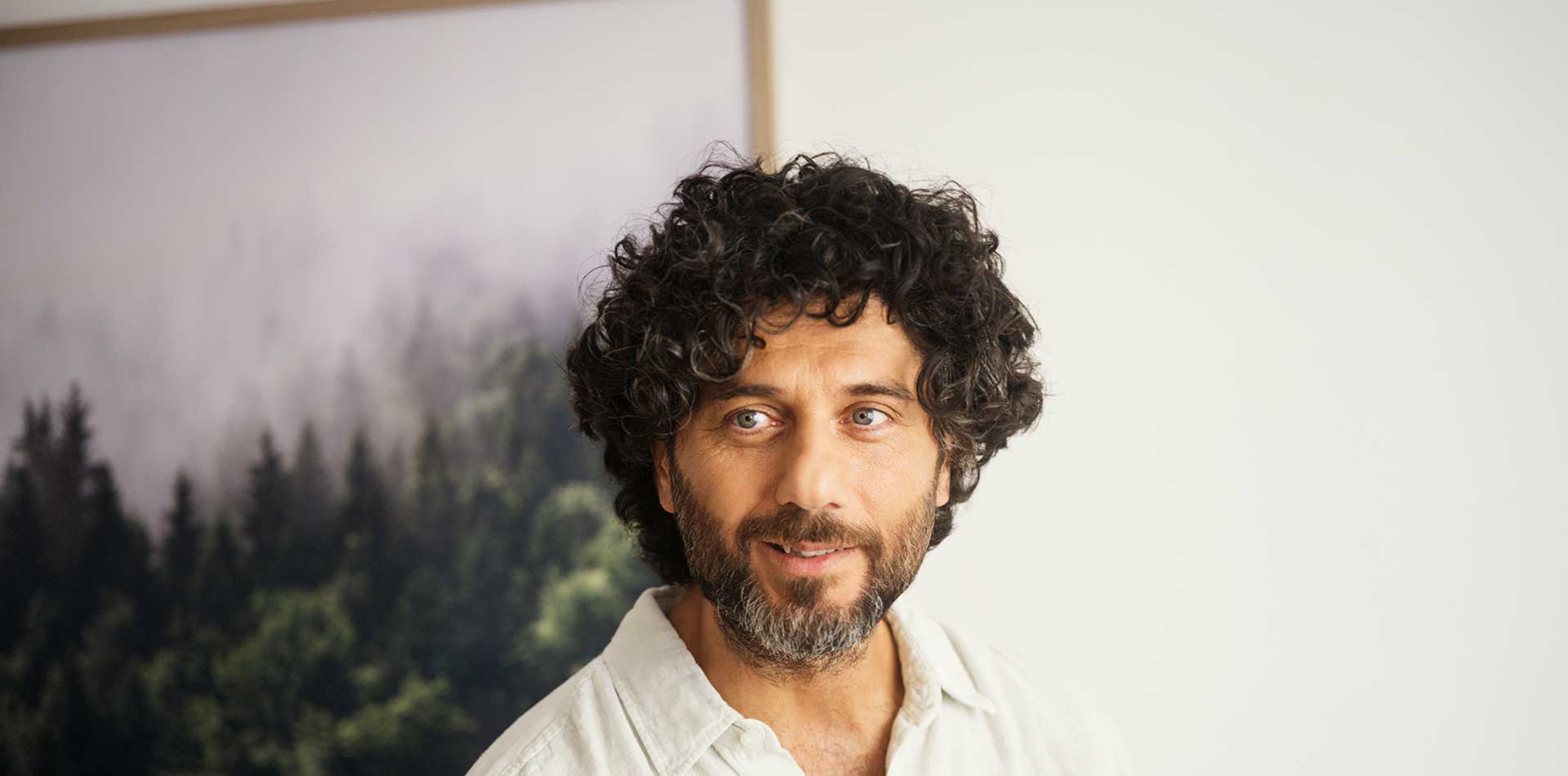 Mies seisoo sisätilassa ja katsoo kameraan, taustalla seinällä metsäaiheinen taulu.