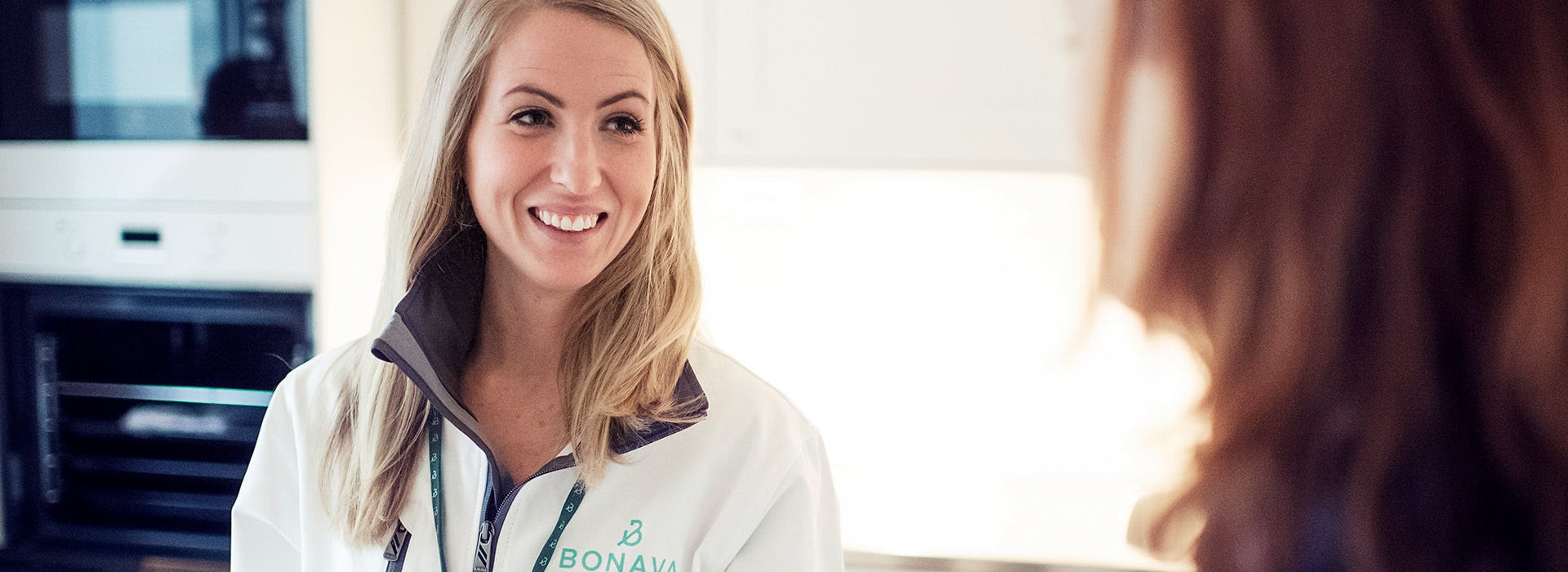 Bonavan asiantuntija hymyilee ja keskustelee asiakkaan kanssa modernissa keittiössä. Ystävällinen kohtaaminen ja asiantunteva palvelu luovat luottamusta uuden kodin hankintaan.