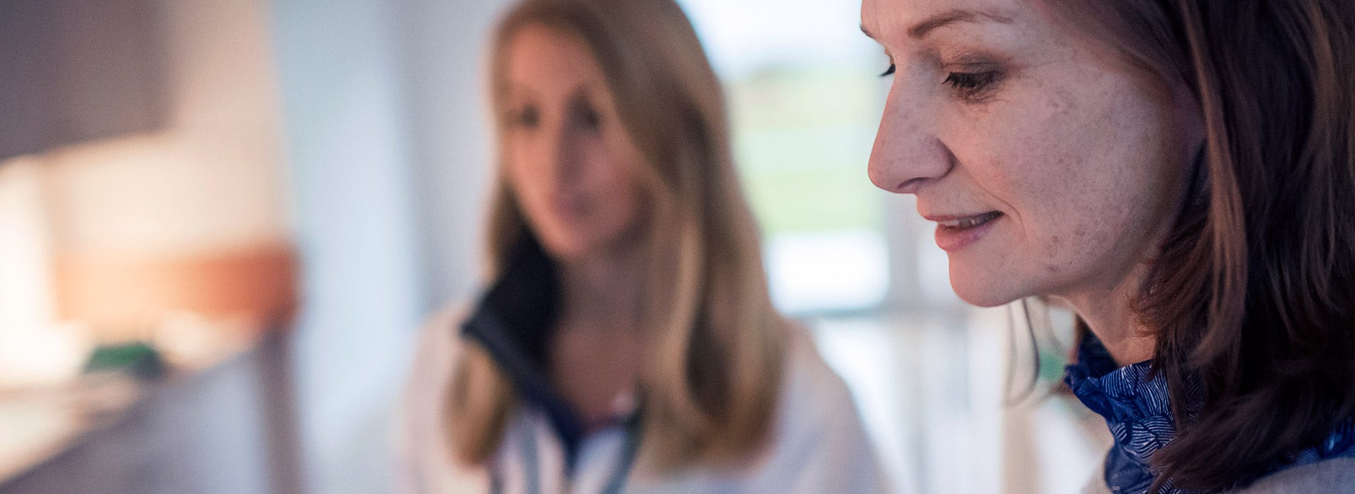 Etualalla hymyilevä nainen sinisessä huivissa, taustalla toinen nainen epätarkkana. Pehmeä valaistus ja lämmin tunnelma viestivät keskittyneestä keskustelusta tai päätöksenteosta kodikkaassa ympäristössä.