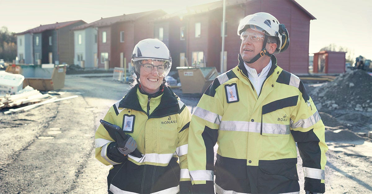 Terveellisen ja turvallisen työpaikan perusta rakentuu välittämisen kulttuurista Kaksi Bonavan työntekijää seisoo aurinkoisella rakennustyömaalla kypärät ja suojalasit päässä. Heillä on yllään keltaiset huomiotakit, ja toinen pitää tablet-laitetta kädessään. Taustalla näkyy punaisia rivitaloja rakenteilla.