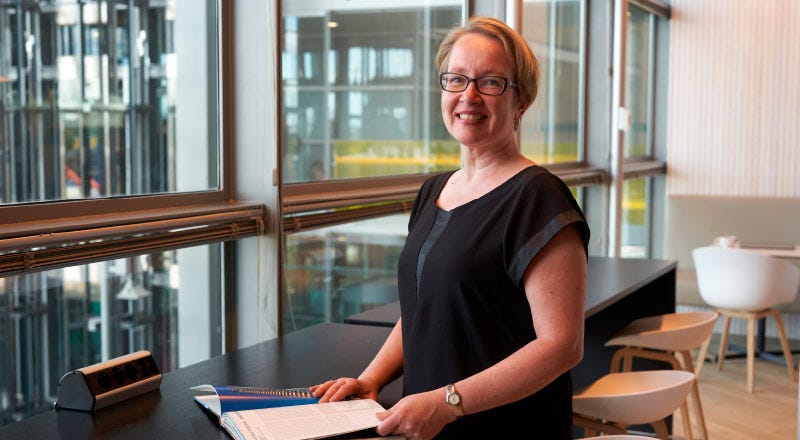 Millainen on hyvä asuntopohja? Projektijohtaja Mari Karttunen seisoo modernissa ja valoisassa toimistotilassa hymyillen. Hän nojaa korkeaan pöytään, jolla on avoin lehti. Taustalla suuret ikkunat, joista avautuu näkymä rakennuksen sisätiloihin ja hissikuiluun.