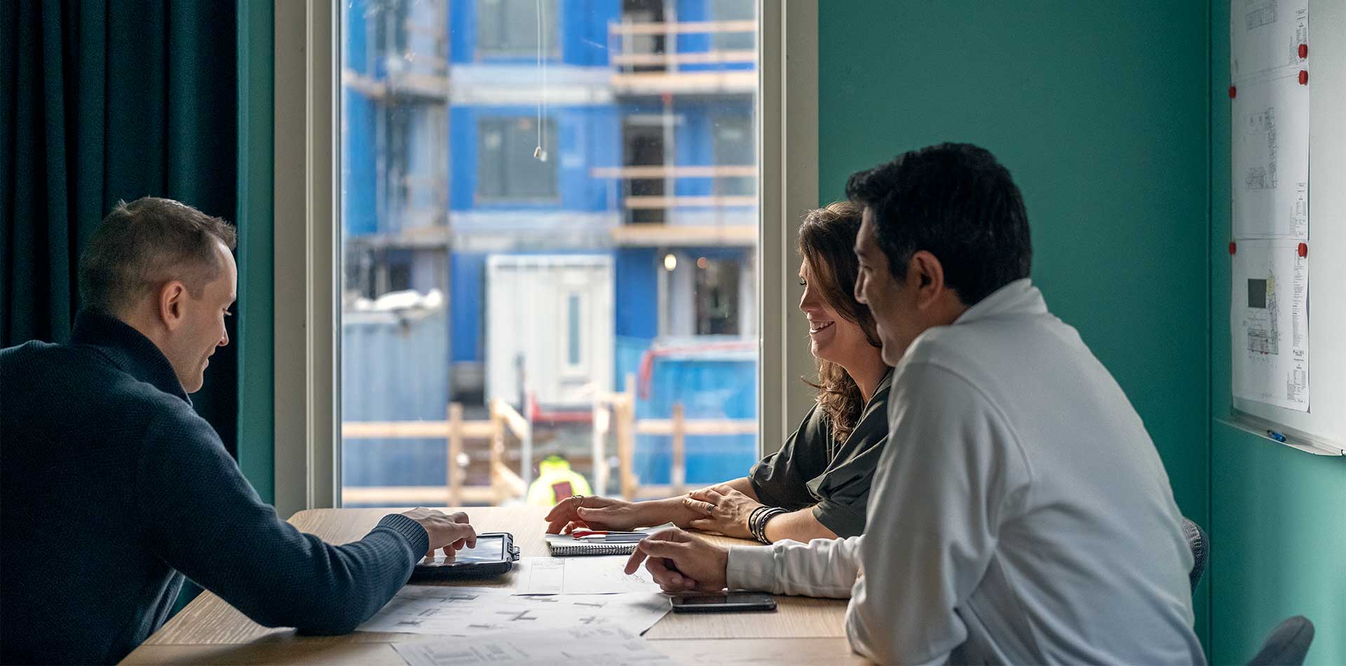 Kolme henkilöä istuu pöydän ääressä keskustelemassa rakentamiseen liittyvässä tapaamisessa. Suunnitelmia näkyy seinällä, ja ikkunasta avautuu näkymä rakennustyömaalle, jossa näkyy parakkeja ja työmaa-aitoja.