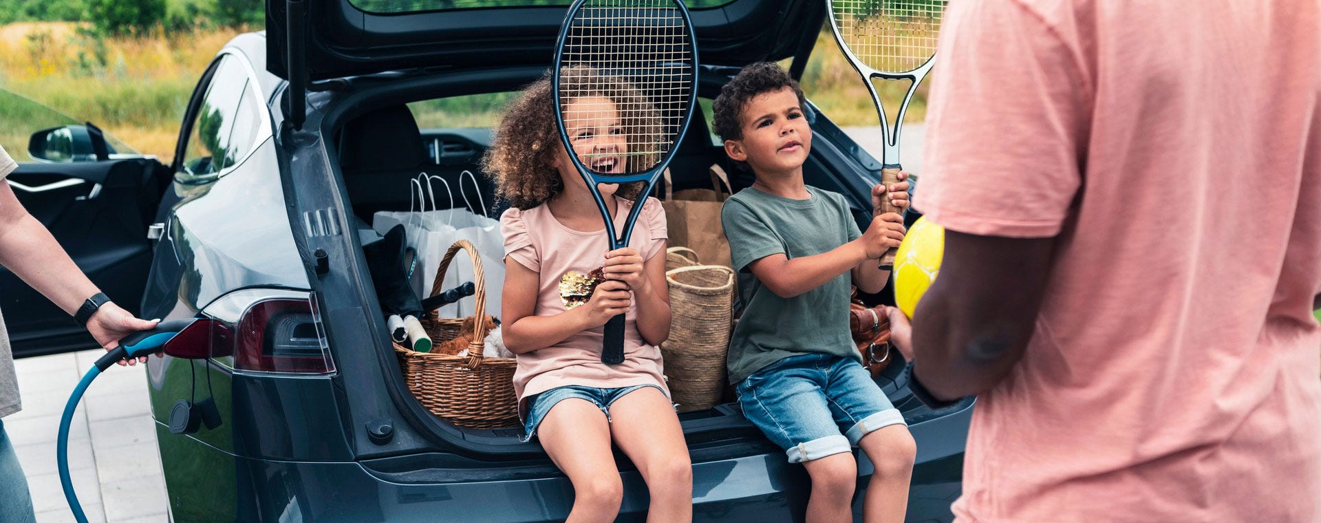 Perhe valmistautuu liikunnalliseen päivään sähköauton latautuessa. Lapset istuvat takaluukun reunalla pidellen tennismailoja, takana kori ja urheilutarvikkeita. Aikuinen pitelee palloa ja keskustelee lasten kanssa.