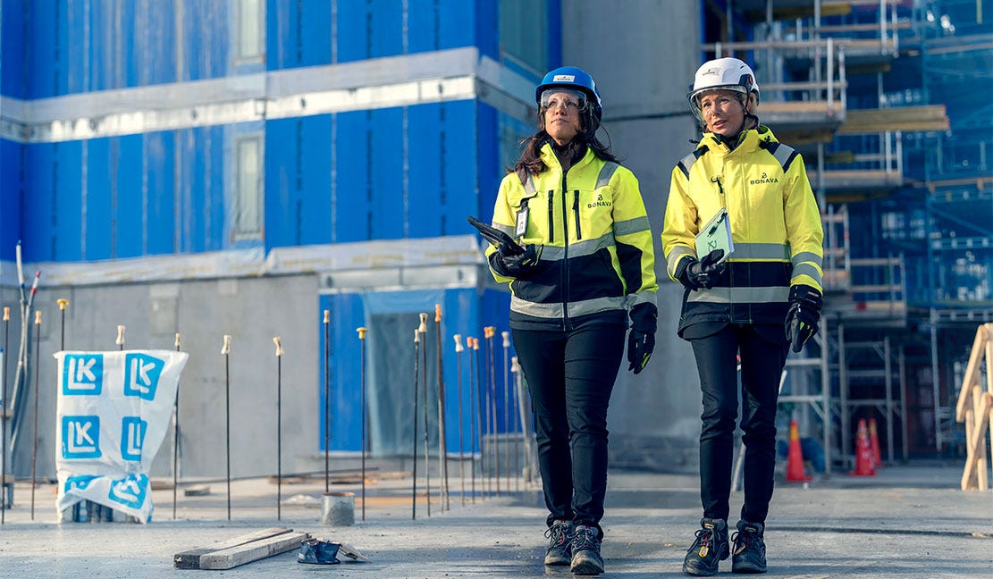Huippupisteet BREEAM-luokituksessa Kaksi rakennusalan ammattilaista kävelee työmaalla kypärät ja suojavaatteet yllään. Heillä on mukanaan tabletit ja asiakirjat. Taustalla näkyy rakenteilla oleva sinisellä suojamuovilla peitetty rakennus ja rakennustelineitä.