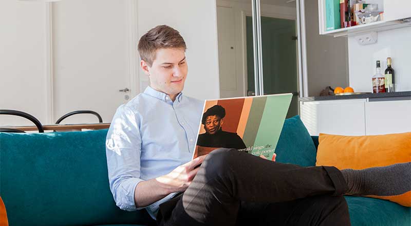 Mies istuu olohuoneen sohvalla ja katsoo vinyylilevyn kantta, taustalla valkoinen keittiö ja värikkäitä tyynyjä sohvalla.
