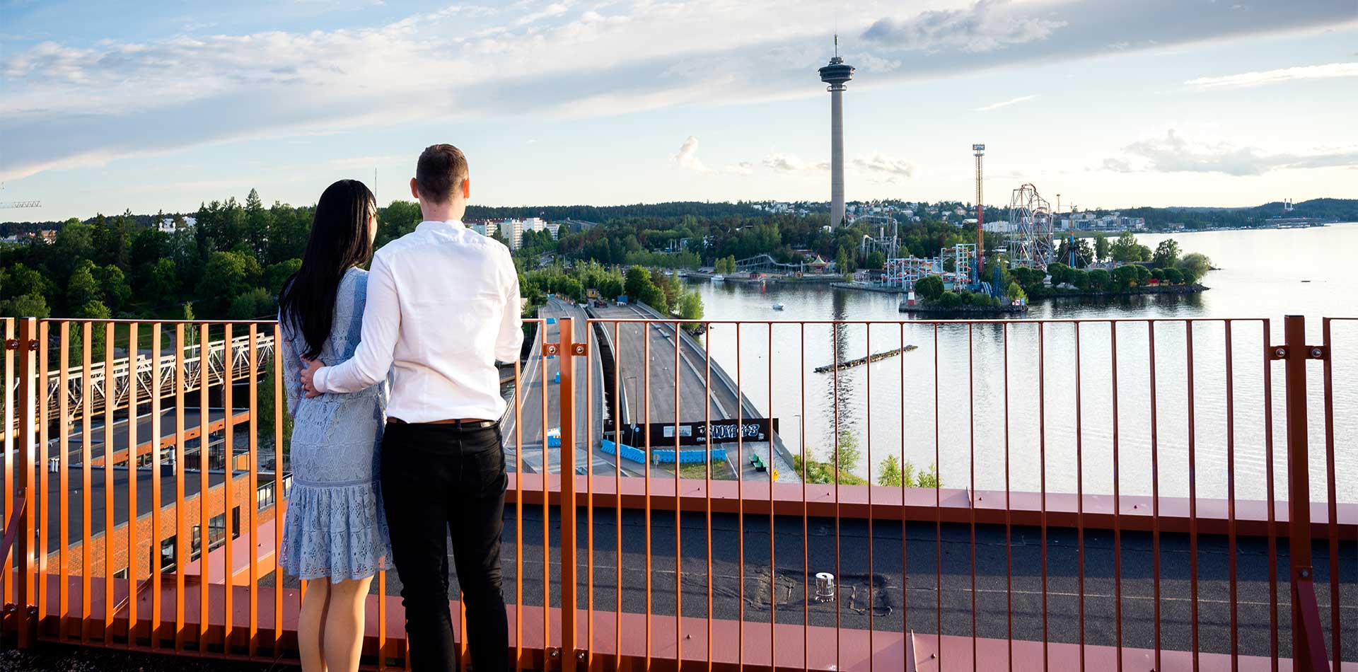 Pariskunta seisoo kattoterassilla katsellen kohti Tampereen Näsijärveä ja Särkänniemen huvipuistoa. Taustalla kohoaa Näsinneula, ja ilta-aurinko valaisee maiseman lämpimästi. 