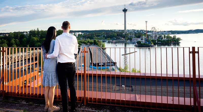 Pariskunta seisoo kattoterassilla katsellen kohti Tampereen Näsijärveä ja Särkänniemen huvipuistoa. Taustalla kohoaa Näsinneula, ja ilta-aurinko valaisee maiseman lämpimästi. 