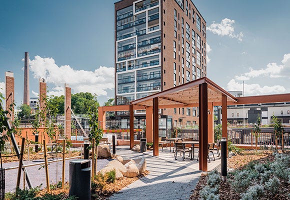 Vehreä sisäpiha, jossa moderni pergola ja viihtyisä oleskelualue. Kivetyt käytävät, istutukset ja luonnonläheinen miljöö luovat rauhallisen ympäristön. Taustalla kohoaa nykyaikainen asuintalo suurine lasipintoineen.