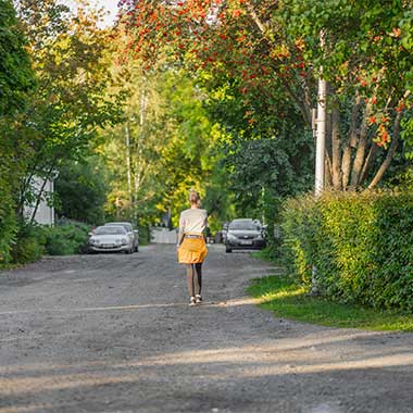 Henkilö kävelee rauhallista puistotietä pitkin vehreässä ympäristössä. Puiden oksilla roikkuvat punaiset marjat tuovat väriä maisemaan. Taustalla näkyy autoja ja omakotitaloja.