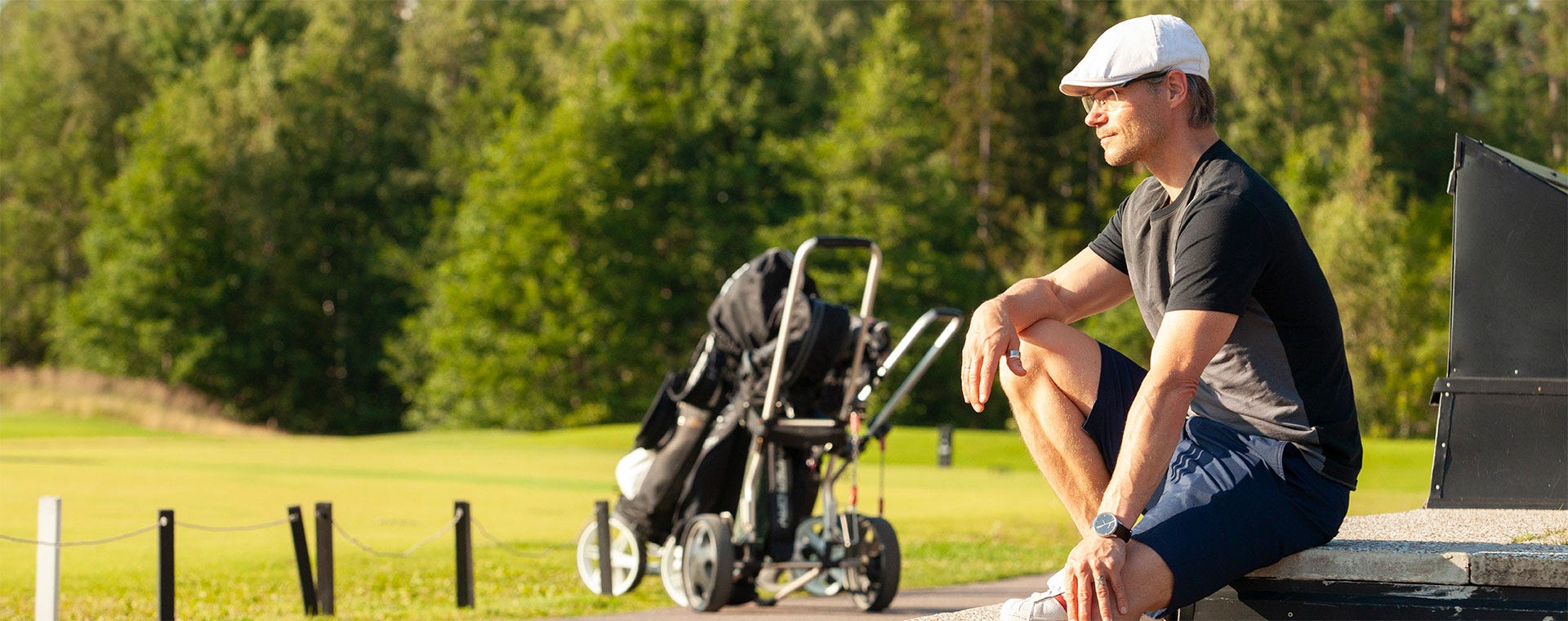 Mies istuu auringonpaisteessa golfkentän laidalla. Hänellä on päällään lippalakki, aurinkolasit ja urheilullinen asu. Vieressä seisoo golfkärry mailoineen. Taustalla vehreä kenttä ja metsän reunustama maisema.