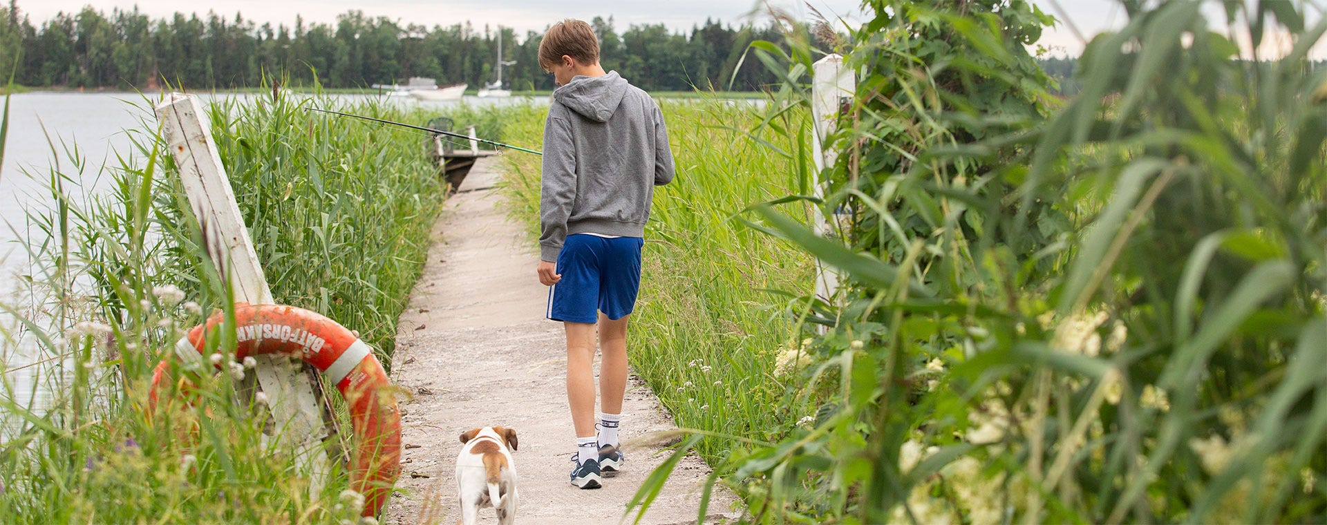 Poika kävelee kapealla laiturilla kohti järveä pieni koira vierellään. Hänellä on yllään harmaa huppari ja siniset shortsit, ja kädessä näkyy onkivapa. Reunustavat vihreät kaislat ja pelastusrenkaan heijastus luovat kesäisen tunnelman.