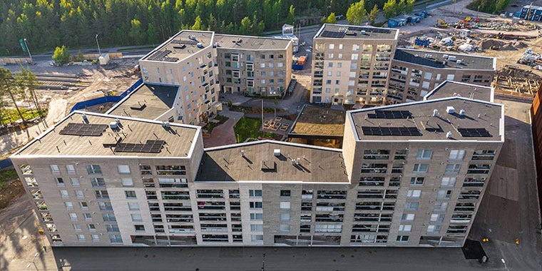 Ilmakuva Postinkantajan kerrostalokorttelista, jossa U-muotoiset rakennukset ympäröivät vehreää sisäpihaa. Rakennusten katoilla on aurinkopaneeleita.
