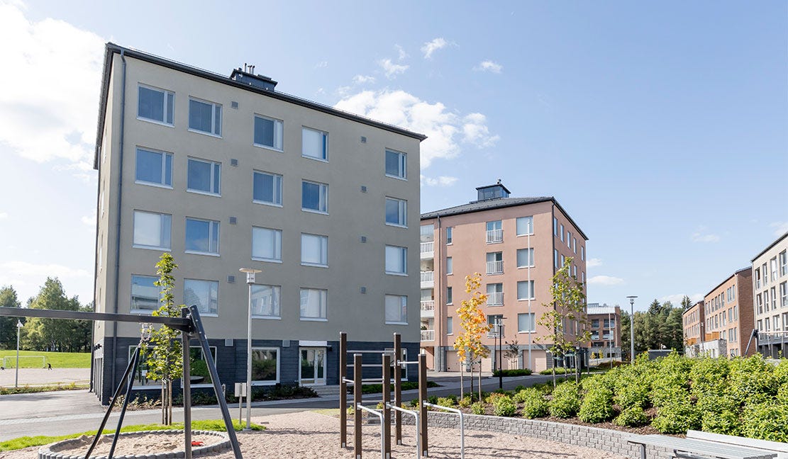Haagan Huvilat, Helsinki Haagan Huvilat - Moderni kerrostaloalue, jossa vaaleasävyisiä rakennuksia, vehreää piha-aluetta ja leikkipaikka. Etualalla kiipeilyteline ja keinu, taustalla sininen taivas ja vehreä maisema.