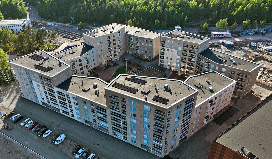 Postinkantaja, Helsinki Helsingin Postinkantaja - Ilmakuva modernista kerrostalokorttelista, jossa U-muotoiset rakennukset ympäröivät vehreää sisäpihaa. Rakennusten katoilla on aurinkopaneeleita. Taustalla näkyy metsää ja rakenteilla olevia alueita.