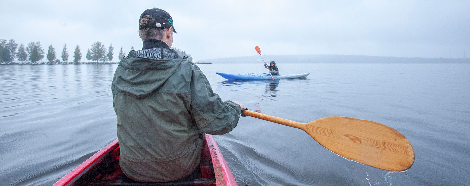 Kaksi melojaa järvellä sumuisena päivänä. Etualalla henkilö soutuveneessä, kädessään puiset airot, ja taustalla toinen meloja sinisessä kajakissa.