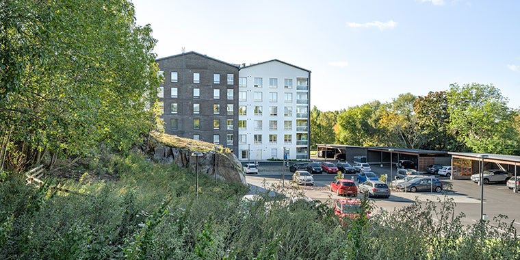 Vähäheikkilän Huvituksen modernit kerrostalot sulautuvat vehreään ympäristöön. Etualalla kallioista maastoa ja vihreää kasvillisuutta, taustalla asukkaiden pysäköintialue katoksineen.