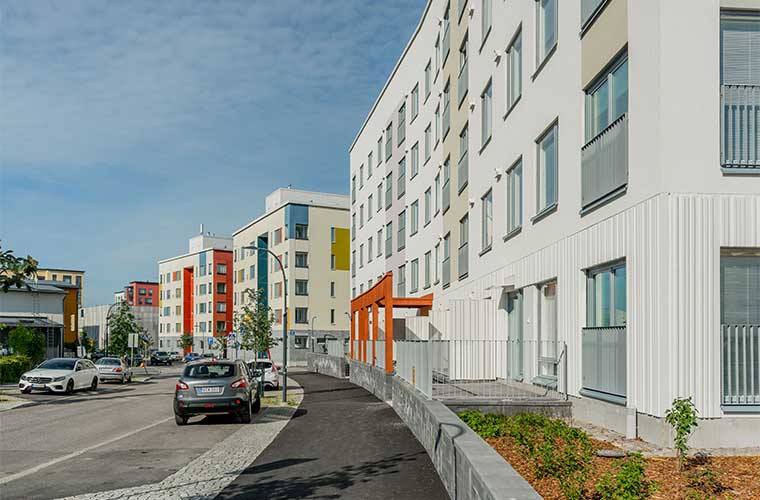 Vantaan Seela 1 -kerrostalo modernissa asuinympäristössä Leinelässä. Rakennuksen vaalea julkisivu yhdistyy värikkäisiin yksityiskohtiin, ja kadun varrella näkyy autoja sekä istutuksia.
