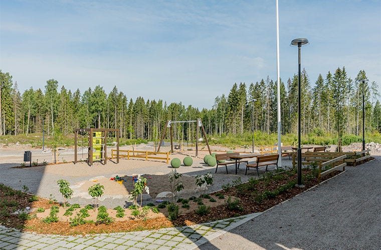 Vantaan Seela 1:n suojaisa pihapiiri, jossa leikkipaikka, keinut, penkkejä ja viheristutuksia. Pihaa ympäröi metsäinen maisema, ja taustalla kohoaa vehreä kuusikko. Hiekkapohjainen alue tarjoaa tilaa ulkoiluun ja rentoutumiseen.