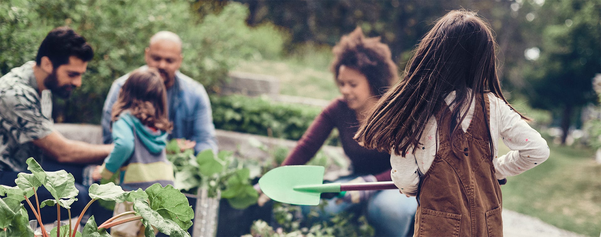 Perhe viettää aikaa yhteisöllisessä kaupunkipuutarhassa. Etualalla lapsi ruskeassa essussa pitää vihreää lapioa, taustalla aikuiset ja toinen lapsi hoitavat kasveja vehreässä ympäristössä.