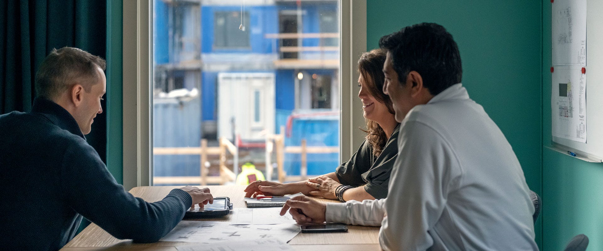 Kolme henkilöä istuu pöydän ääressä keskustelemassa rakentamiseen liittyvässä tapaamisessa. Suunnitelmia näkyy seinällä, ja ikkunasta avautuu näkymä rakennustyömaalle, jossa näkyy parakkeja ja työmaa-aitoja.