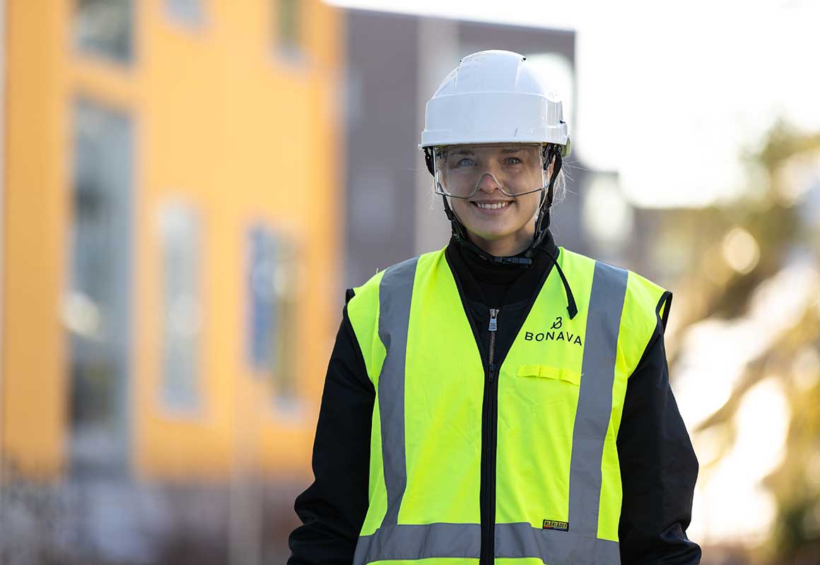 Bonavan työntekijä seisoo rakennustyömaalla päällään kypärä, suojalasit ja huomioliivi, taustalla asuinrakennuksia.