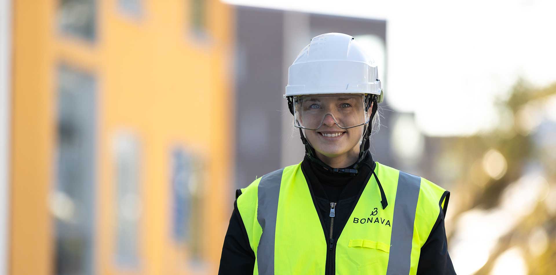 Bonavan työntekijä seisoo rakennustyömaalla päällään kypärä, suojalasit ja huomioliivi, taustalla asuinrakennuksia.