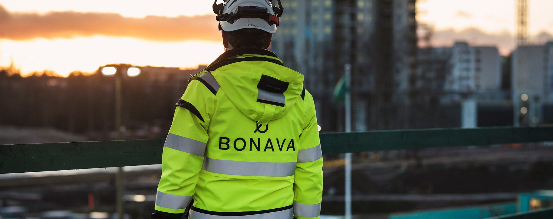 Rakennustyöntekijä seisoo työmaalla auringonlaskun aikaan selkä kameraan päin. Hänellä on yllään keltainen Bonava-logolla varustettu huomiotakki ja suojakypärä. Taustalla näkyy kaupunkimaisema ja rakenteilla olevia kerrostaloja.
