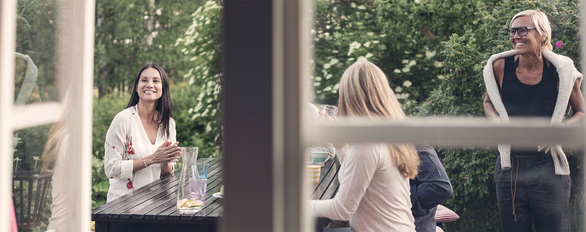 Rentoa yhdessäoloa vehreällä pihalla. Hymyileviä ihmisiä istuu ulkopöydän äärellä ja keskustelee rennosti, samalla kun yksi henkilö saapuu liittymään seuraan.