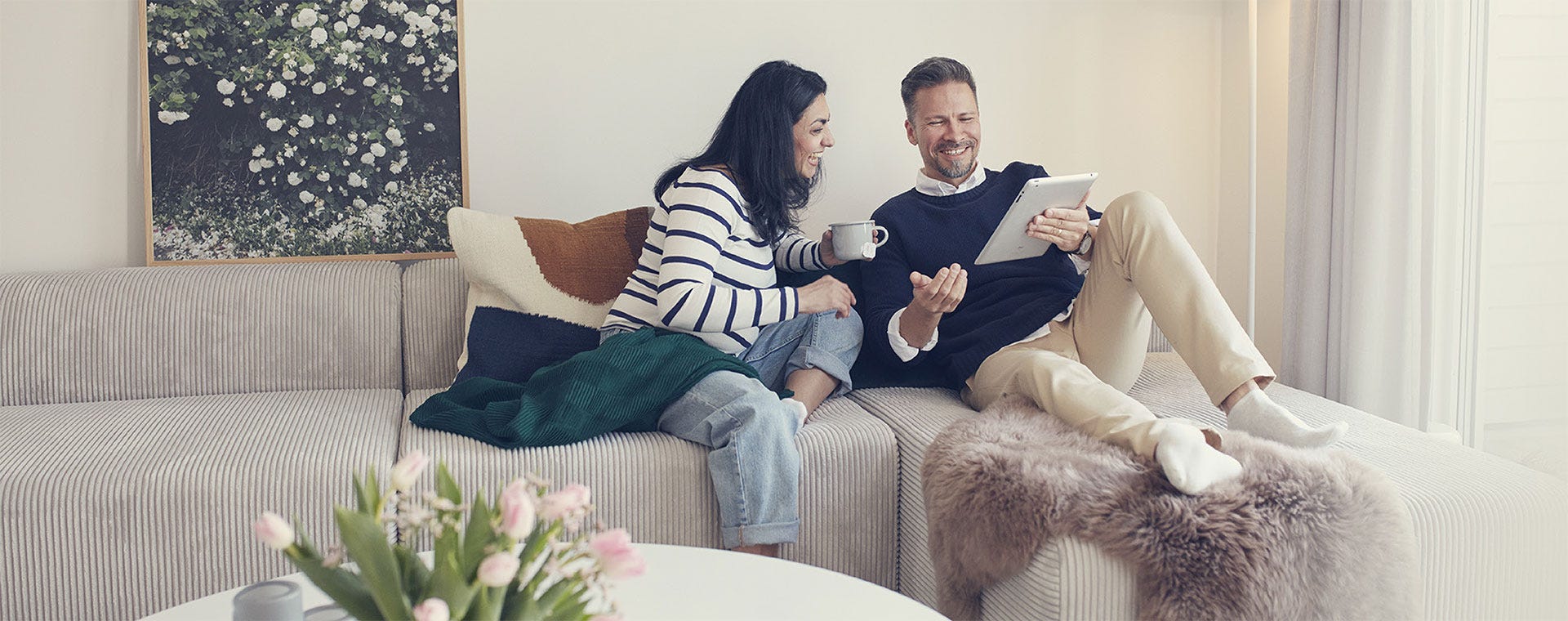 Hymyilevä pariskunta rentoutuu olohuoneen sohvalla. Nainen pitää käsissään teekuppia, ja mies näyttää hänelle jotain tabletin näytöltä. Kodikas tunnelma syntyy lämpimistä sävyistä, sisustustyynyistä ja viltistä.