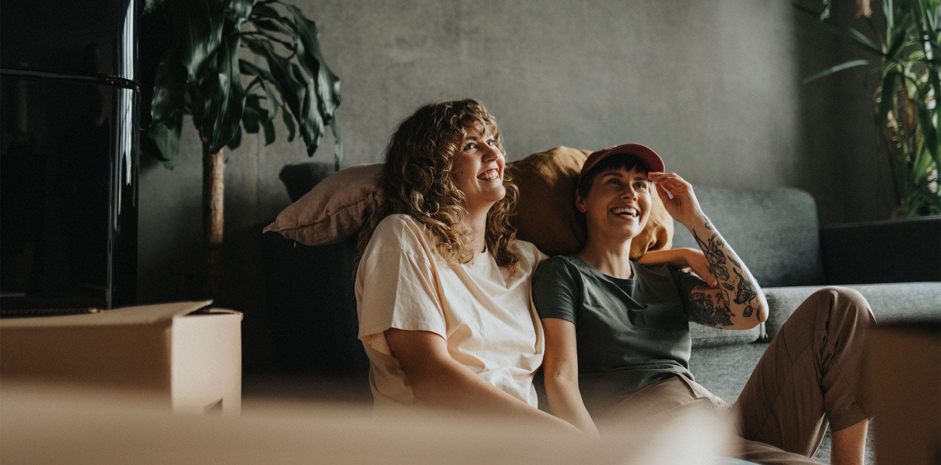 Kaksi henkilöä istuu lattialla sohvaa vasten sisätilassa, ympärillä muuttolaatikoita, ja he katsovat hymyillen eteenpäin.