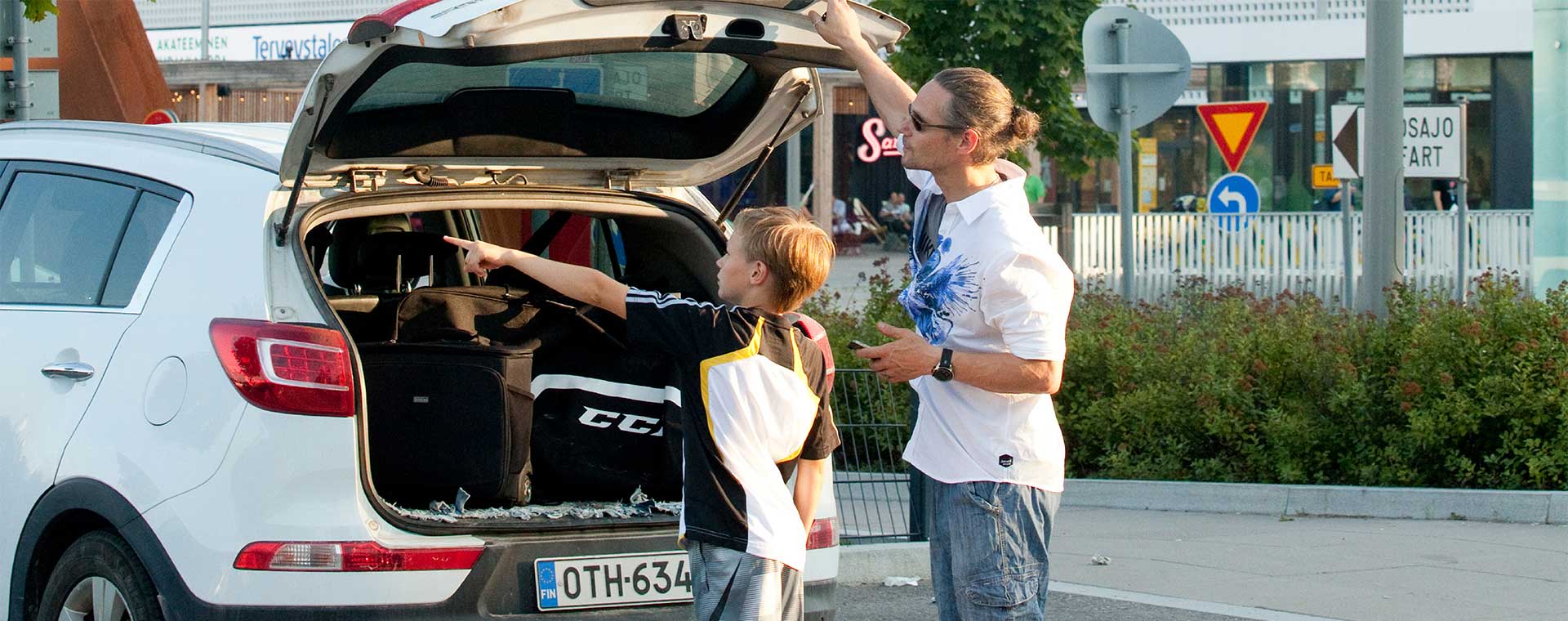 Isä ja poika purkavat auton tavaratilaa kaupunkiympäristössä. Poika osoittaa jotakin tavaroiden joukossa, ja isä kuuntelee häntä, samalla pitäen auton takaluukkua auki. Takana näkyy liikennemerkkejä ja kauppakeskus.