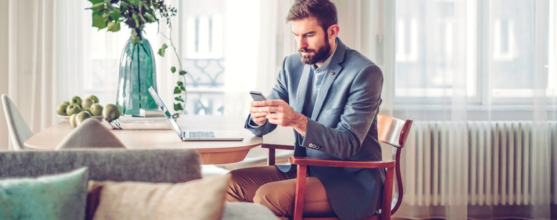 Mies istuu ruokapöydän ääressä tyylikkäässä olohuoneessa, puku päällään ja katsoo keskittyneenä älypuhelintaan. Pöydällä on kannettava tietokone, vihreä maljakko ja kulhollinen omenoita.