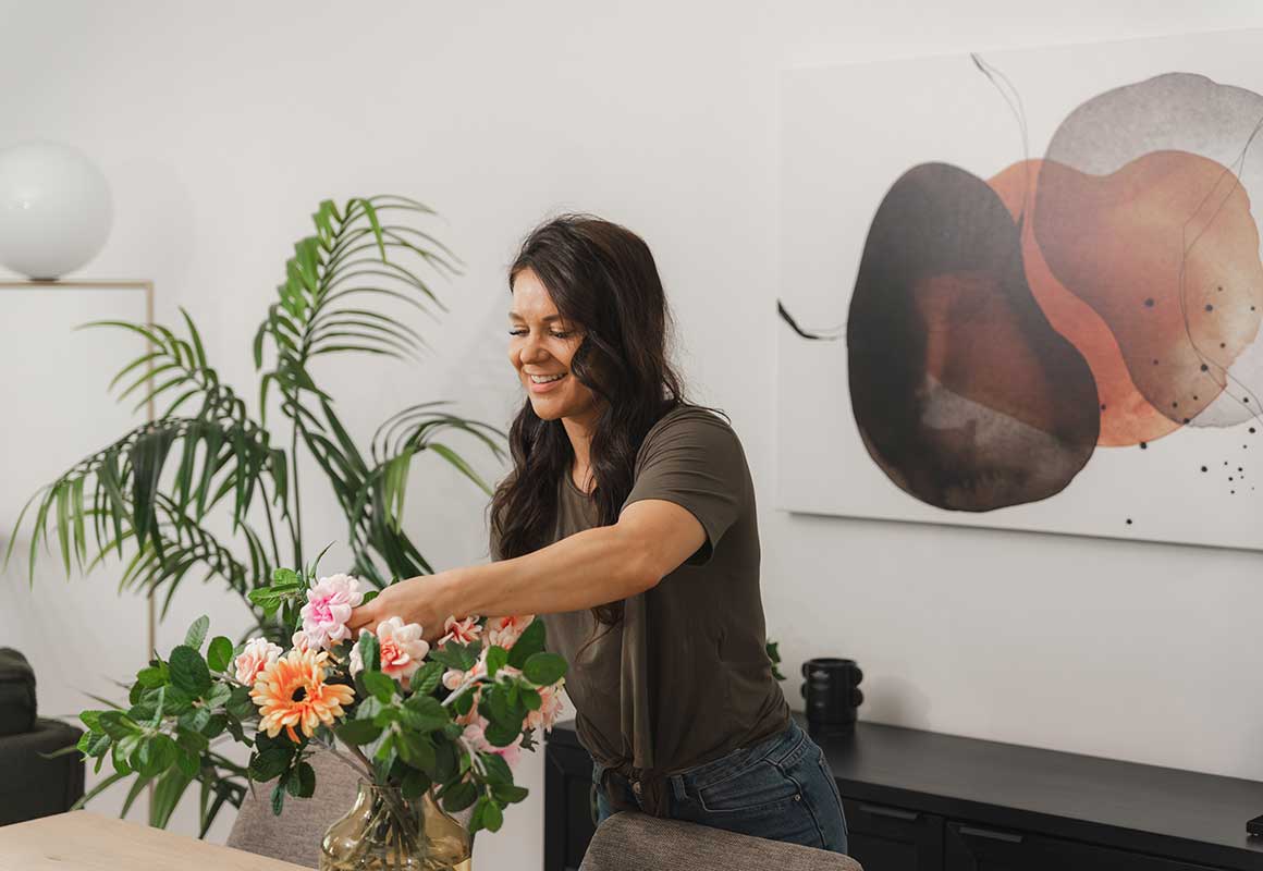 Nainen asettelee kukkia maljakkoon sisätilassa, taustalla huonekaluja ja seinätaulu.