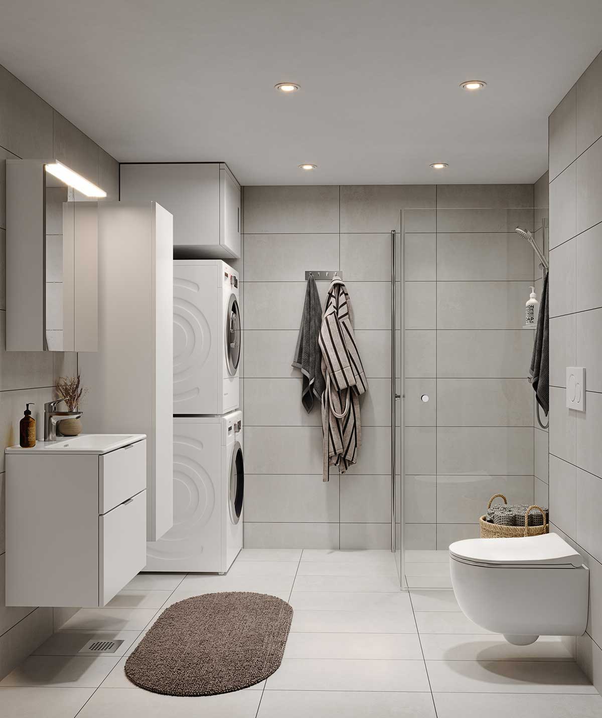 Tyylikäs ja rauhallinen kylpyhuone, jossa on harmaan sävyiset lattia- ja seinälaatat. Valkoiset kylpyhuonekalusteet, kuten allaslaatikosto, pyykkikaappi ja peilikaappi, tuovat valoisuutta tilaan. Seinään kiinnitetty wc-istuin ja lasiseinällä erotettu suihkutila lisäävät modernia ilmettä. Lattialla ovaalinmuotoinen ruskea kylpyhuonematto tuo lämpöä sisustukseen.
