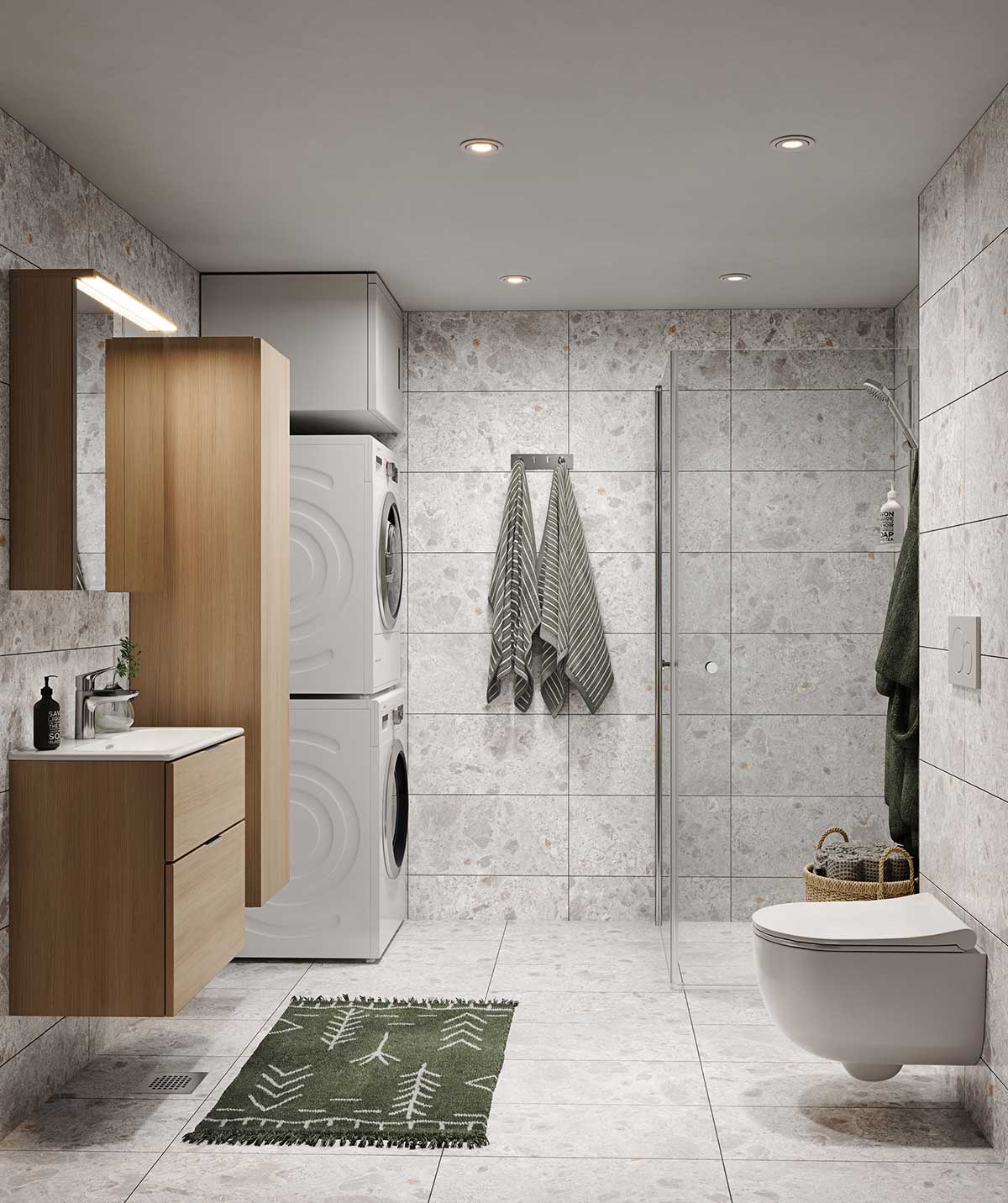Kylpyhuone, jossa harmaan sävyiset, kivikuvioidut lattia- ja seinälaatat tuovat luonnonläheistä tunnelmaa. Tamminen allaskaappi ja peilikaappi tuovat lämpöä tilaan. Seinään kiinnitetty wc-istuin ja lasiseinällä erotettu suihkutila lisäävät modernia ilmettä. Pesukone ja kuivausrumpu on sijoitettu päällekkäin, säästäen tilaa. Vihreä, kuusikuvioitu kylpyhuonematto ja tummanvihreät pyyhkeet tuovat kontrastia ja luonnonläheisyyttä sisustukseen.