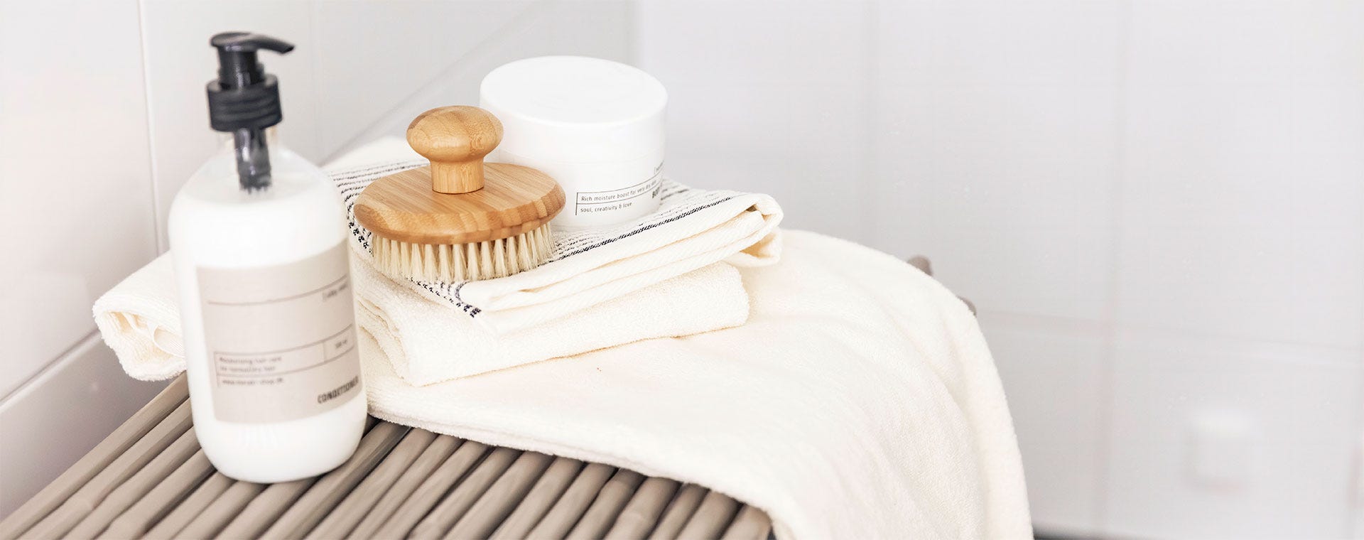 Kylpyhuonetarvikkeita vaalealla penkillä. Pehmeitä valkoisia pyyhkeitä on aseteltu siististi, niiden päällä luonnonmateriaalinen vartaloharja ja tyylikkäitä purkkeja ihonhoitotuotteille. Vasemmalla valkoinen pumppupullo.