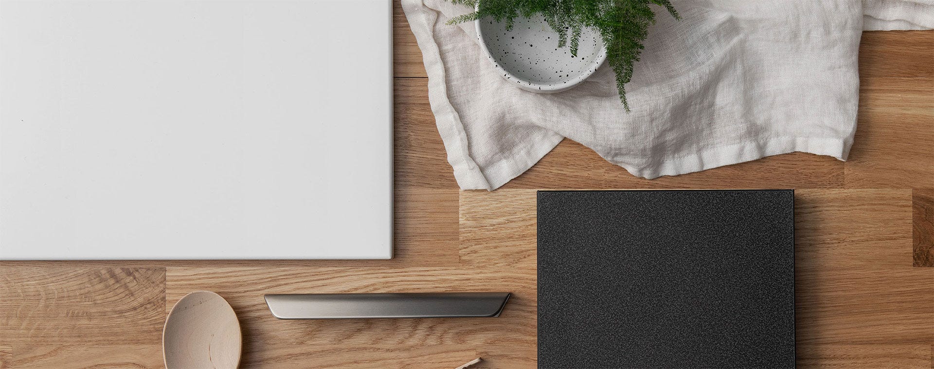Keittiön materiaalinäytteet aseteltuna puiselle pinnalle. Valkoinen ja musta laattapinta, metallinen vetokahva sekä luonnonmateriaalista valmistettu lusikka ja kangasservietti. Pieni ruukkukasvi tuo raikkaan vihreän sävyn kokonaisuuteen.
