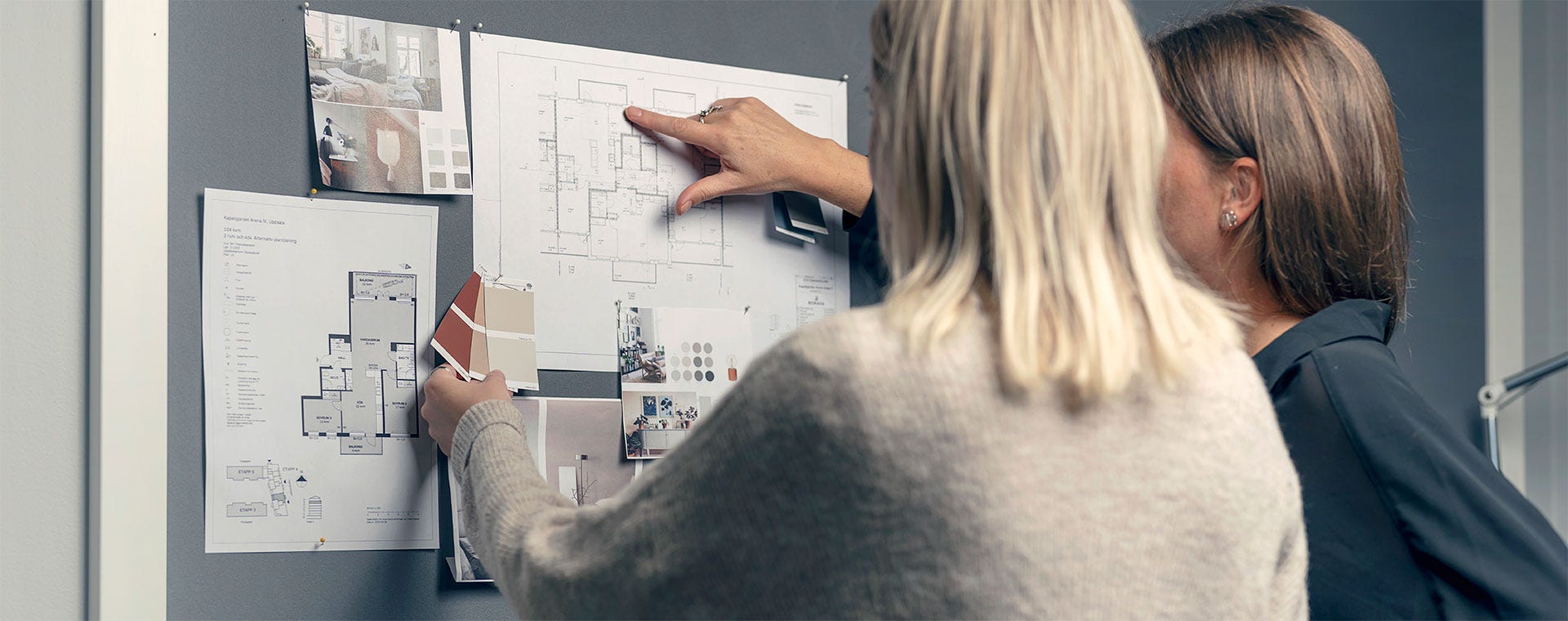 Kaksi naista suunnittelee sisustusta katsellen seinälle kiinnitettyjä pohjapiirustuksia, värimalleja ja inspiraatiokuvia. Toinen osoittaa sormellaan pohjapiirrosta, kun taas toinen pitelee värikortteja. Taustalla näkyy tunnelmallisia sisustuskuvia ja huolella valittuja materiaalinäytteitä.