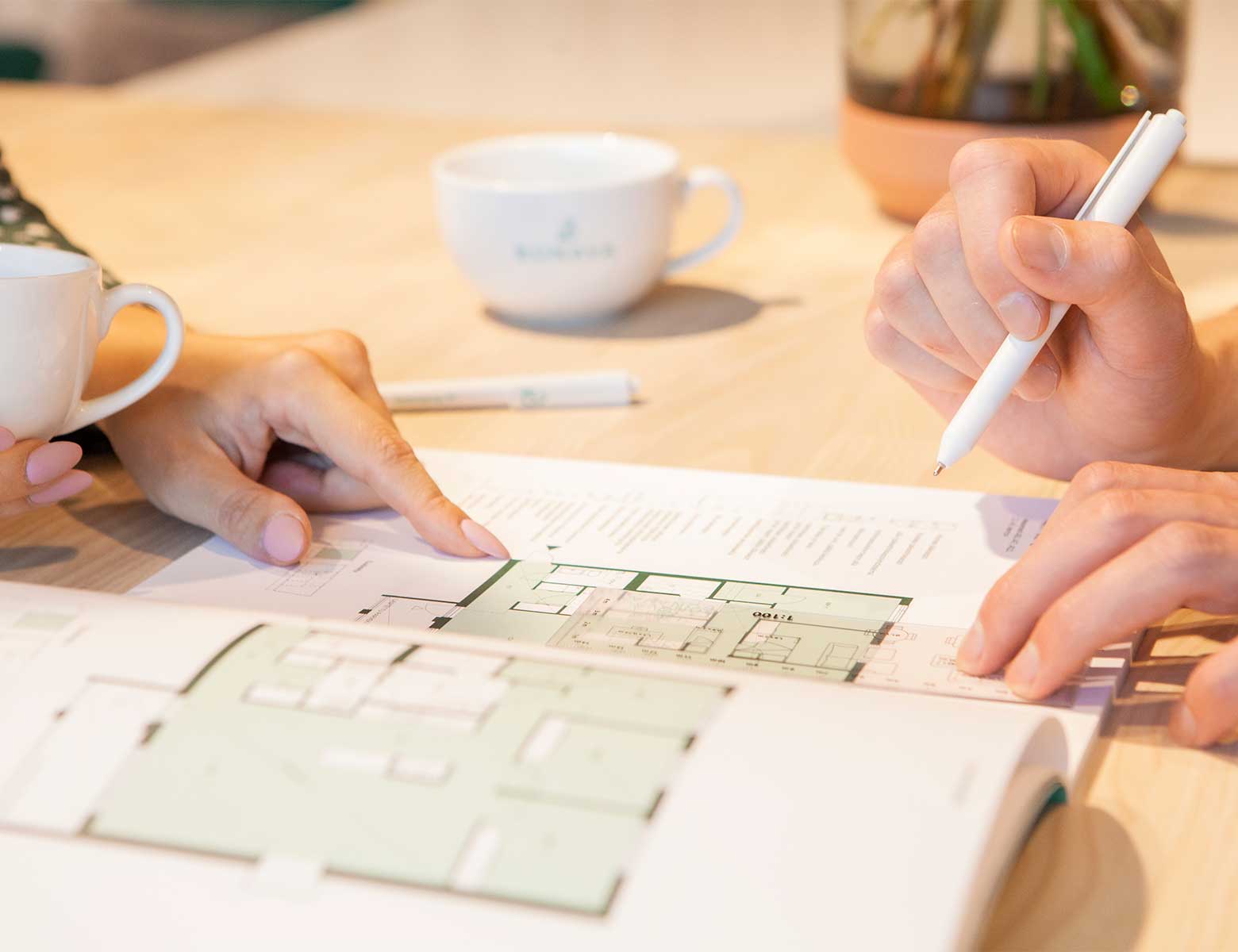 Kaksi henkilöä tarkastelee asunnon pohjapiirrosta avonaisessa esitteessä. Toinen osoittaa sormellaan yksityiskohtaa pohjakuvassa, kun taas toinen pitelee kynää merkintöjen tekemistä varten. Pöydällä on kahvikuppeja ja kasvi, luoden rennon mutta keskittyneen tunnelman asuntohankinnan tai -sijoittamisen suunnitteluun.