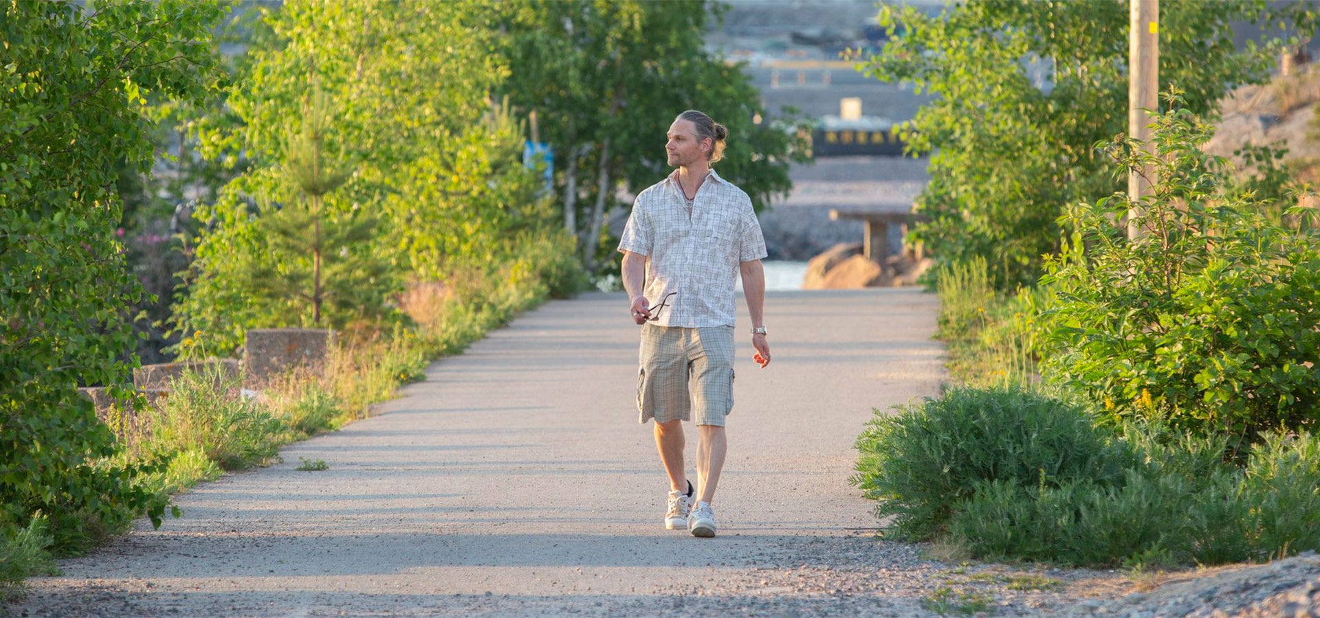 Mies kävelee kesäisessä maisemassa vehreällä puistotiellä ilta-auringossa.