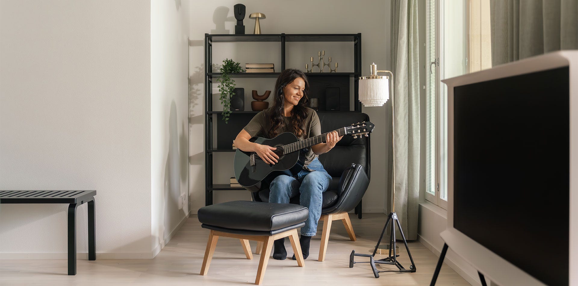 Nainen istuu modernissa olohuoneessa mustalla nojatuolilla ja soittaa akustista kitaraa hymyillen. Taustalla on hyllykkö sisustusesineineen, ja luonnonvalo tulvii sisään suurista ikkunoista. 