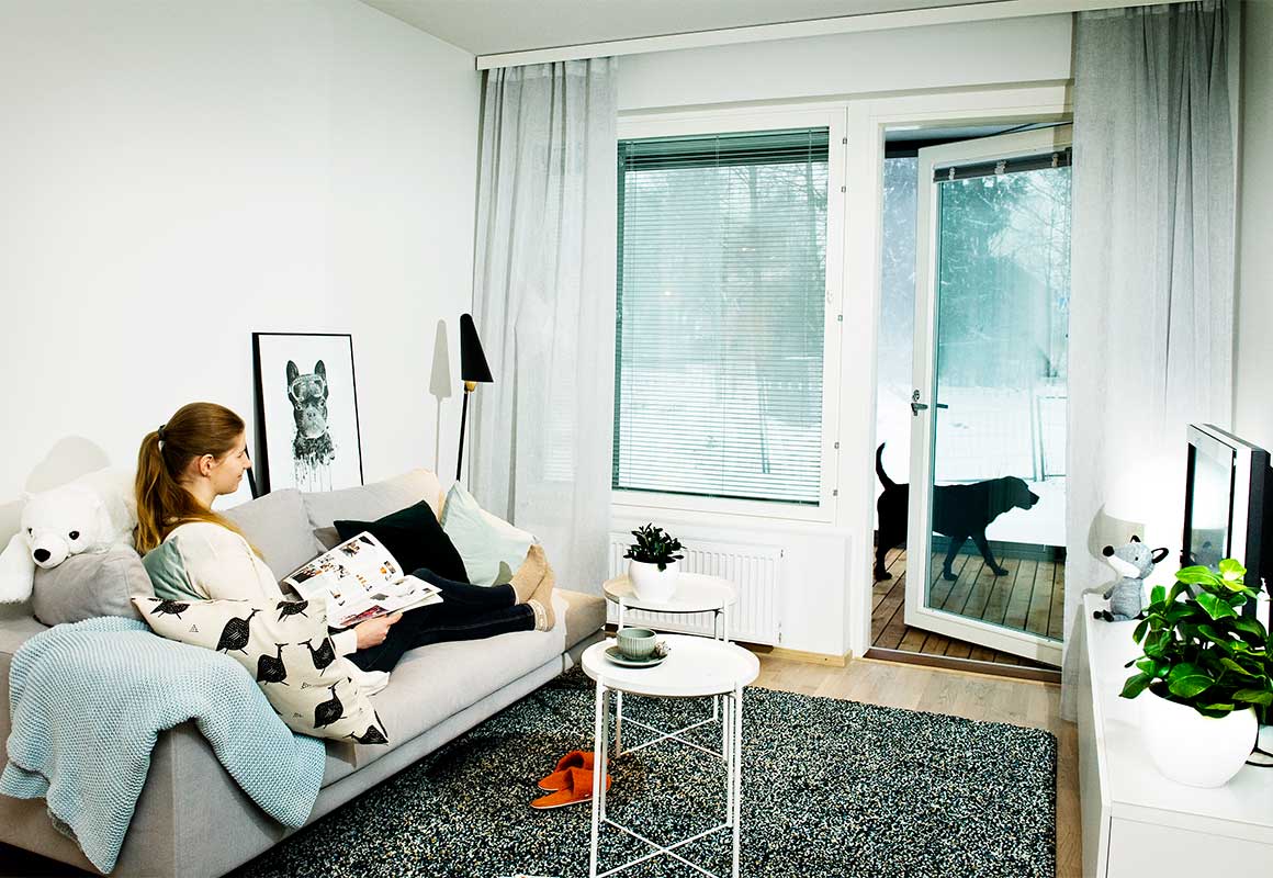 Nainen istuu olohuoneen sohvalla lukemassa lehteä, taustalla suuri ikkuna ja lasiovi, jonka takana parvekkeella seisoo koira.