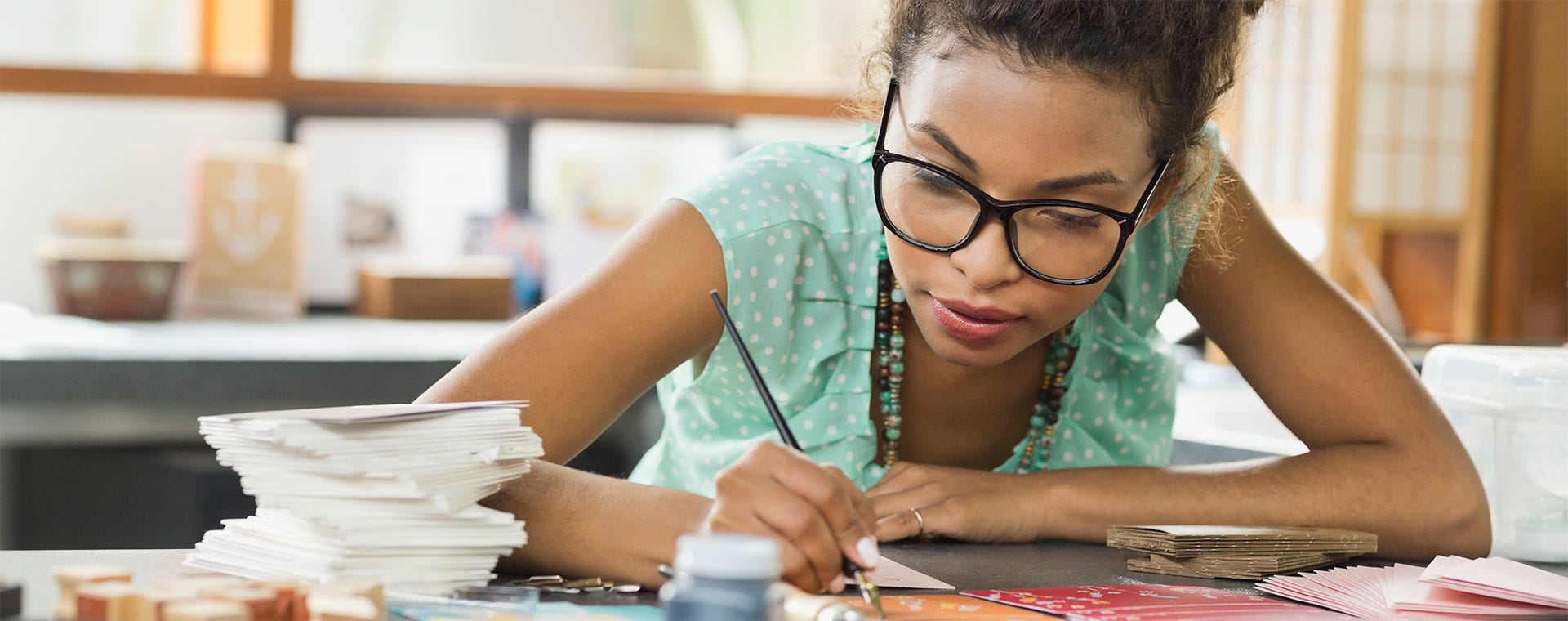  Nuori nainen työskentelee kotona askarteluprojektin parissa, ympärillään pinoja paperia ja askartelutarvikkeita. Keskittynyt ilme ja rauhallinen ympäristö heijastavat kodin tarjoamaa luovuuden ja rauhan tilaa.