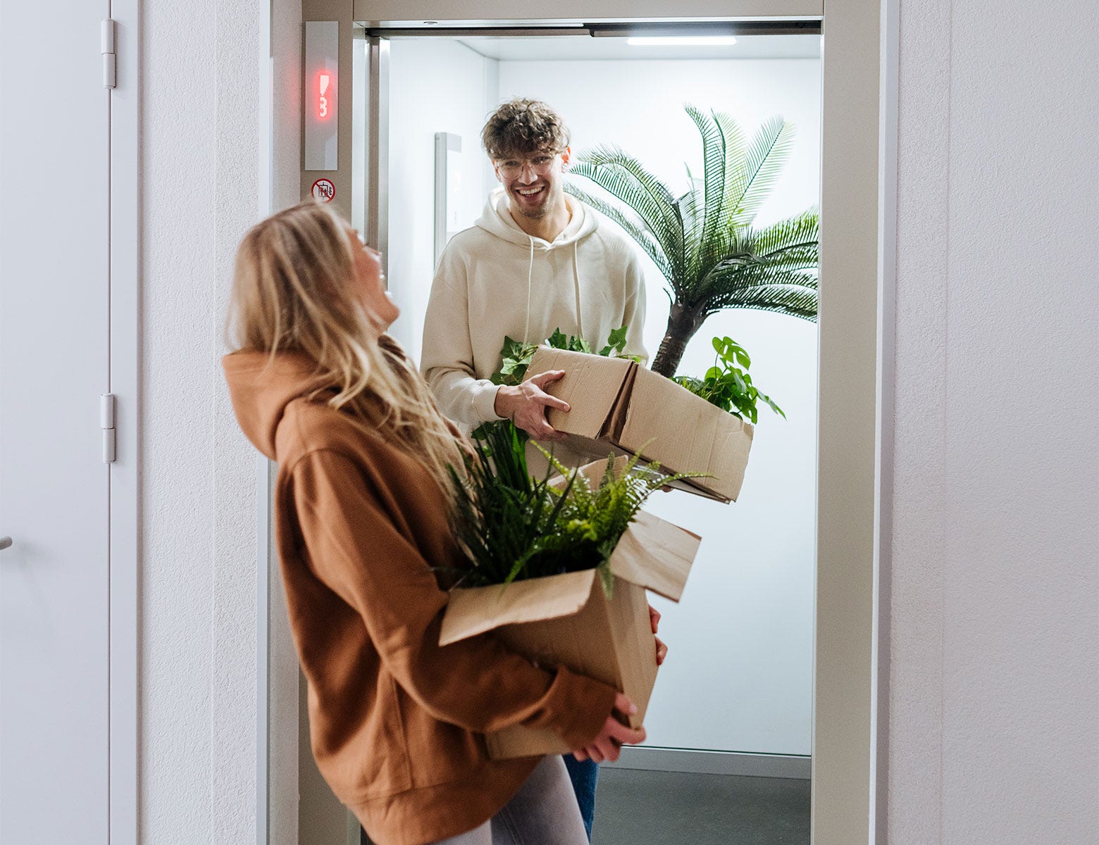 Nuori pariskunta muuttaa uuteen kotiin ja kantaa pahvilaatikoita, joissa on viherkasveja. Huppariin pukeutunut mies seisoo hississä nauraen, kun nainen kantaa laatikoita käytävällä. Muuton ilo ja uuden kodin odotus näkyvät heidän kasvoillaan.