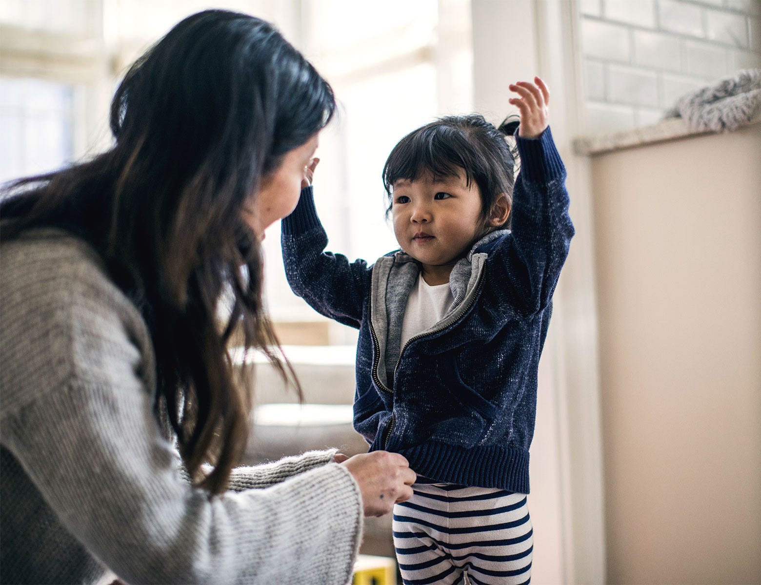 Äiti ja pieni lapsi jakavat iloisen hetken kotona. Lapsi seisoo kädet kohotettuina ja hymyilee, kun äiti katsoo häntä lämpimästi. Taustalla näkyy kodin sisustusta, kuten kaide ja ikkunasta tulviva luonnonvalo, joka luo pehmeän ja kodikkaan tunnelman.