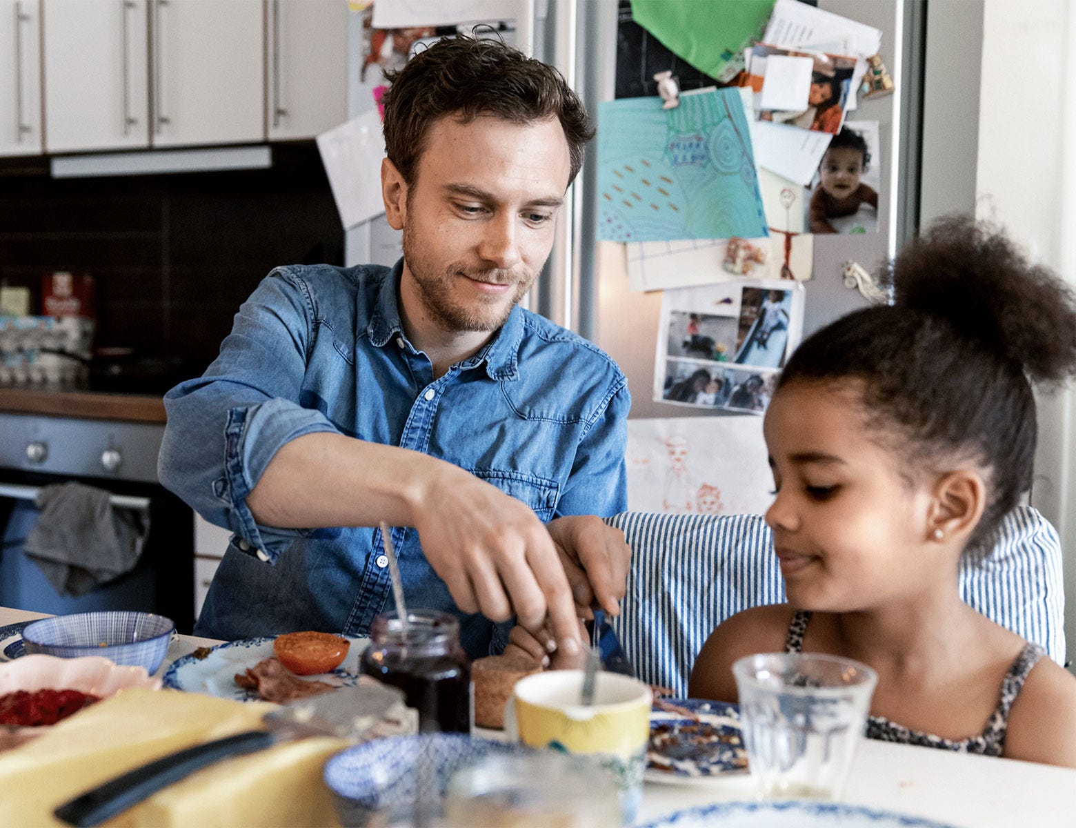 Isä ja tytär syövät aamupalaa keittiössä, jossa jääkaapin ovessa on lasten piirustuksia ja perhekuvia. Isä hymyilee ja valmistaa leipää tyttärelleen, joka katsoo häntä iloisesti. Rentoa perhe-elämää kodikkaassa ympäristössä.