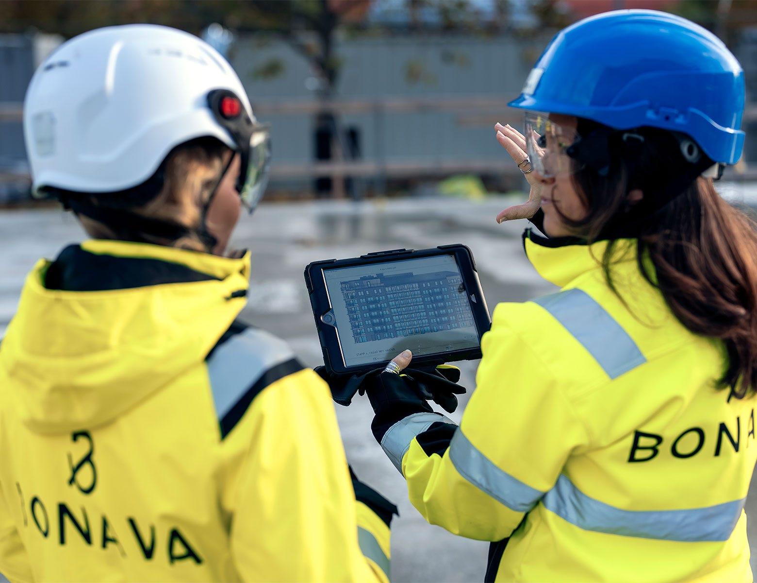  Kaksi rakennustyömaalla työskentelevää henkilöä keltaisissa huomiotakeissa ja kypärissä keskustelee. Toinen heistä pitää kädessään tablettia ja osoittaa näyttöä samalla selittäen. Taustalla näkyy rakennustyömaan aita ja rakenteilla olevia kohteita.