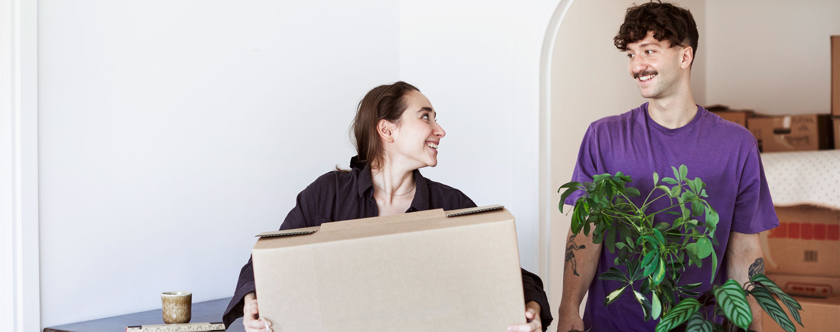 Hymyilevä pariskunta kantaa muuttolaatikoita ja viherkasveja uuteen kotiinsa. Taustalla näkyy muita pakkauslaatikoita, jotka odottavat purkamista. Kuvasta välittyy lämmin ja iloinen tunnelma uuden kodin kynnyksellä.