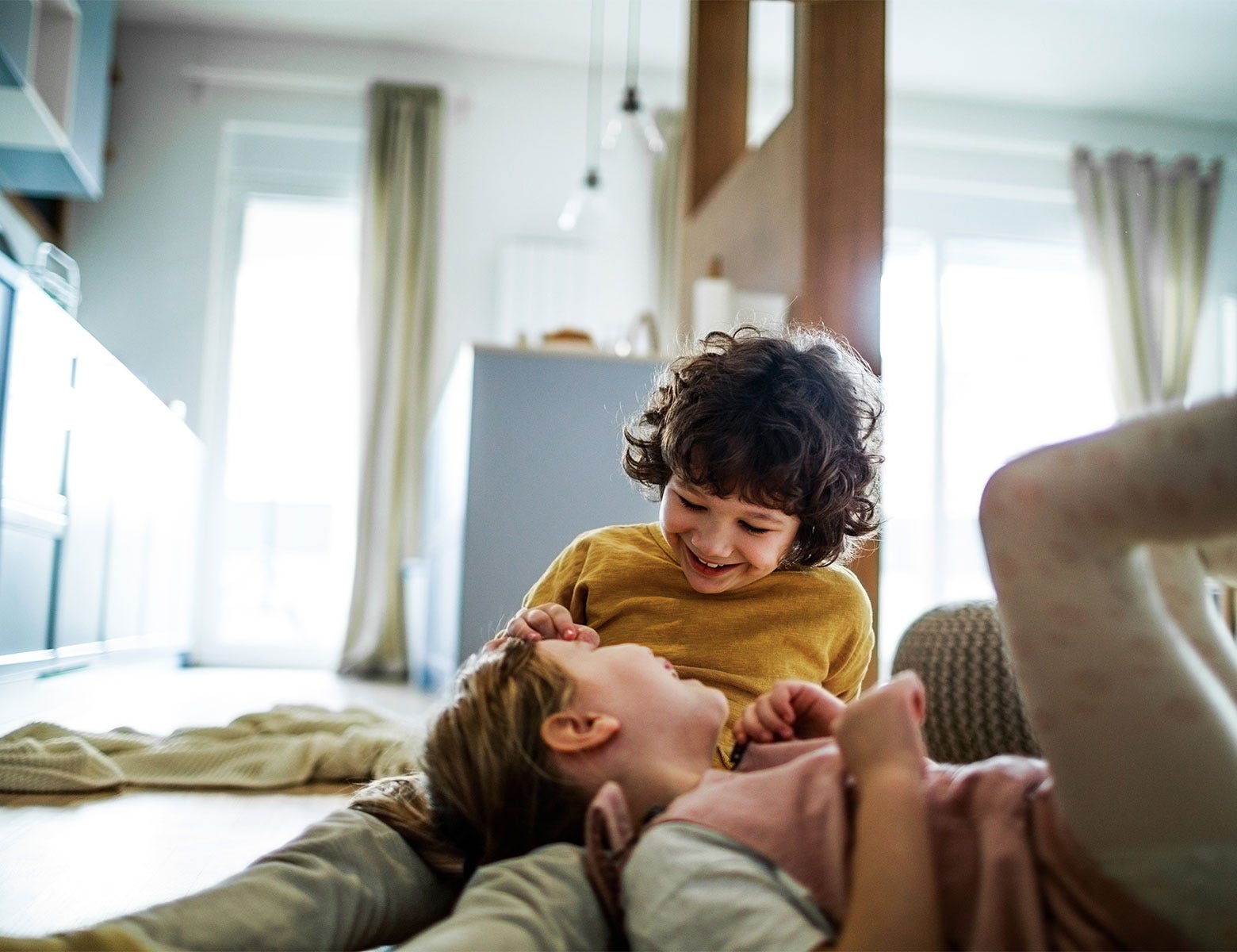  Kaksi lasta leikkii nauraen lattialla kodikkaassa ja valoisassa olohuoneessa. Suuret ikkunat päästävät sisään luonnonvaloa, ja taustalla näkyy keittiö sekä vaaleat verhot, jotka kehystävät tilaa.