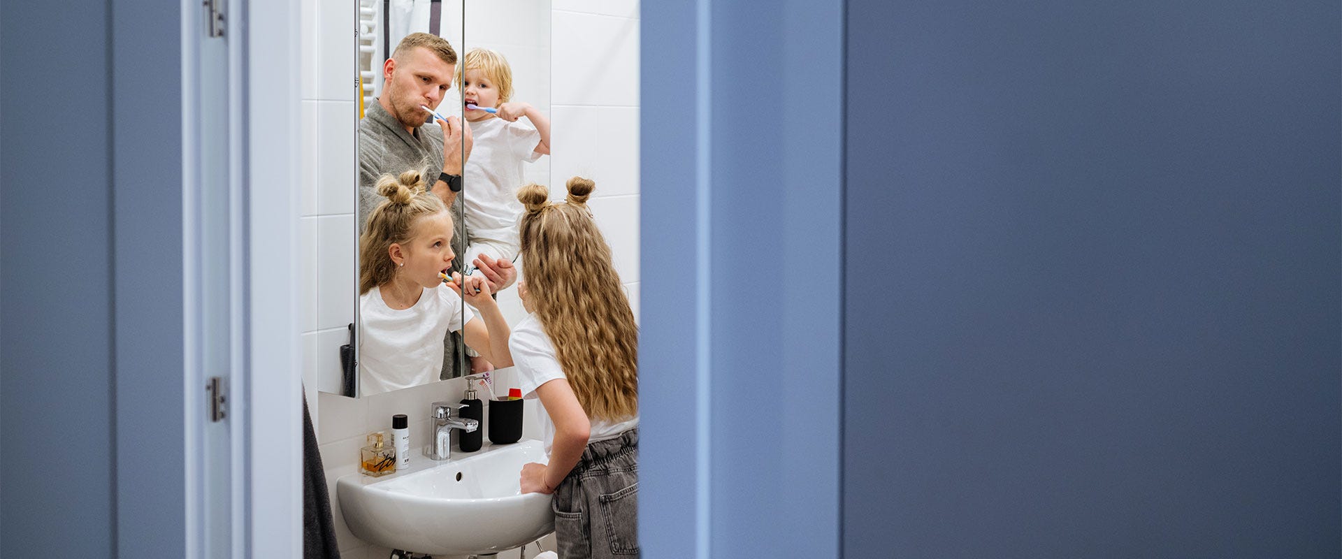 Isä harjaa hampaitaan kylpyhuoneessa kahden lapsensa kanssa, molemmat lapset ovat pukeutuneet valkoisiin t-paitoihin ja harjaavat myös hampaitaan. Peilin kautta näkyvä lämmin perhehetki heijastaa arjen rutiinien turvallisuutta kodin seinien sisällä.